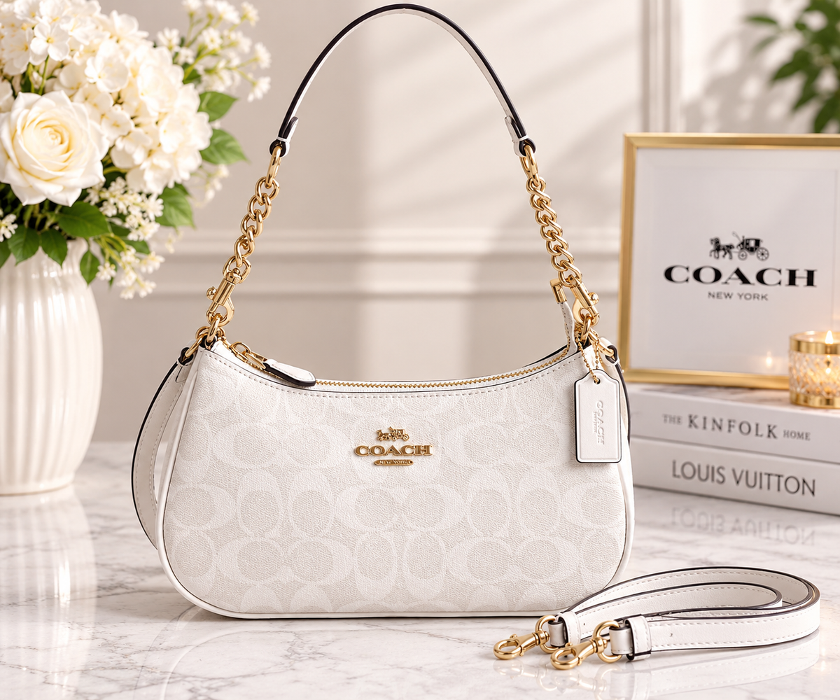 A Coach handbag in white monogram canvas with gold-tone chain straps and hardware, displayed on a marble surface with flowers and branded books in the background.