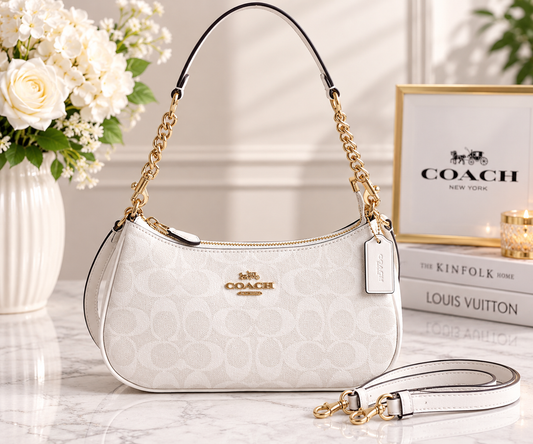 A Coach handbag in white monogram canvas with gold-tone chain straps and hardware, displayed on a marble surface with flowers and branded books in the background.