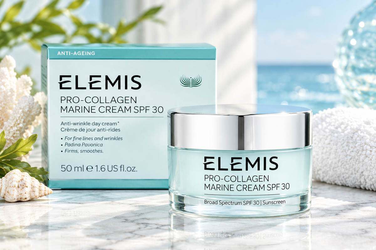 Elemis Pro-Collagen Marine Cream SPF 30 shown with its light blue and white product box and matching jar with a silver lid, set on a marble surface with a blurred beach and seashell background.
