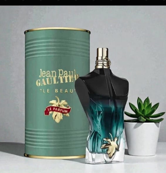 Jean Paul Gaultier Le Beau Le Parfum perfume bottle with a black and teal gradient body, gold cap, and a matching metallic teal cylindrical box with gold lettering, styled on a white surface with a small potted succulent against a gray background.