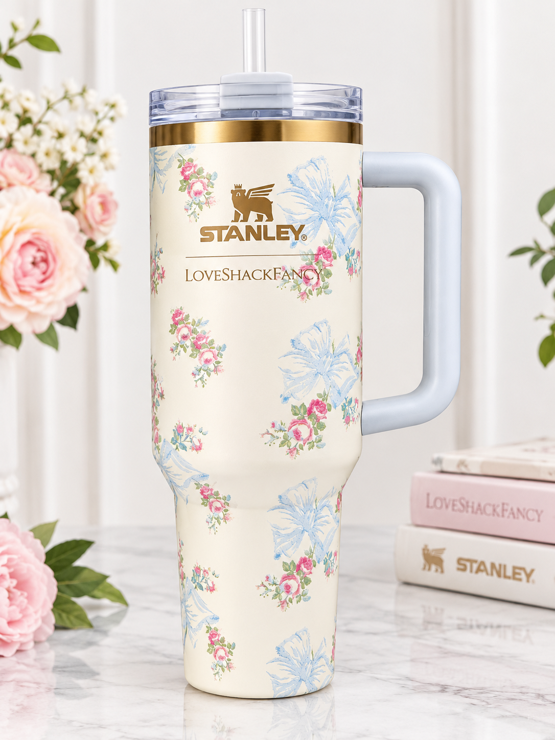 A Stanley insulated tumbler with a floral print, light blue handle, and clear lid with straw, displayed on a marble surface with pink flowers and books in the background.
