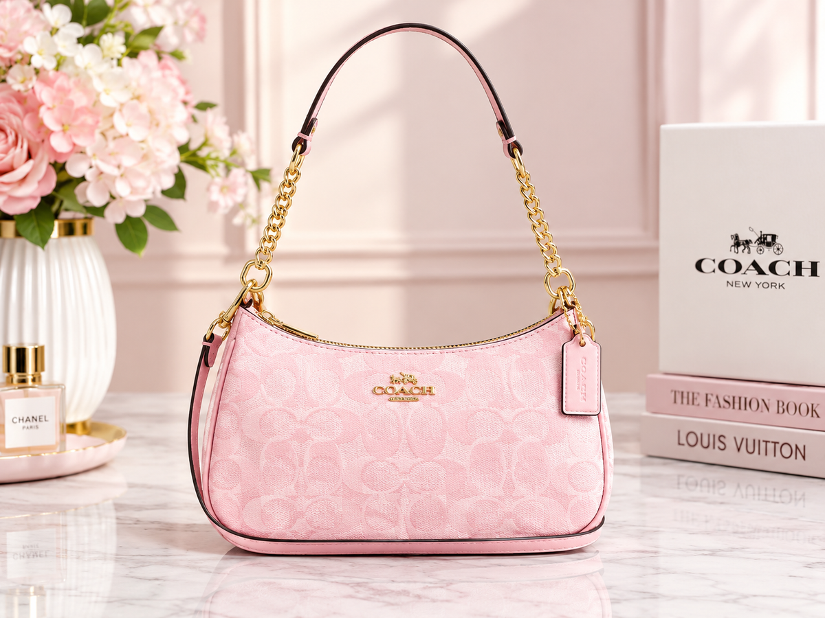A pink Coach shoulder bag with gold-tone chain strap and gold logo detail, displayed on a marble surface with flowers, a perfume bottle, and a Coach box in the background.